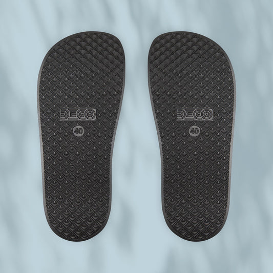 Flat lay view of black slide soles showing tread pattern and branding, on a pale blue background
