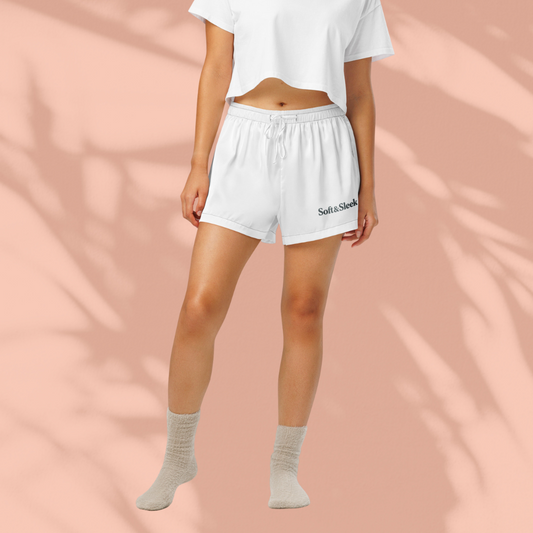 Full-body view of a woman wearing white Soft & Sleek shorts and a matching cropped top, peach background with soft shadows.