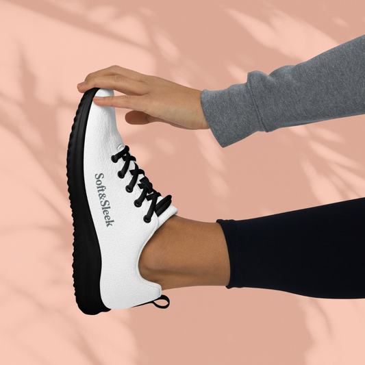 Side view of a person holding their foot up, wearing a white Soft & Sleek sneaker with black sole and black laces.