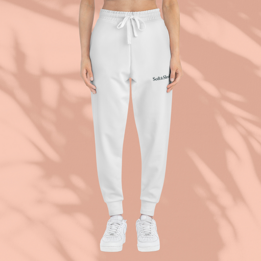 Front view of woman wearing white Soft & Sleek joggers and white sneakers, standing against a peach background