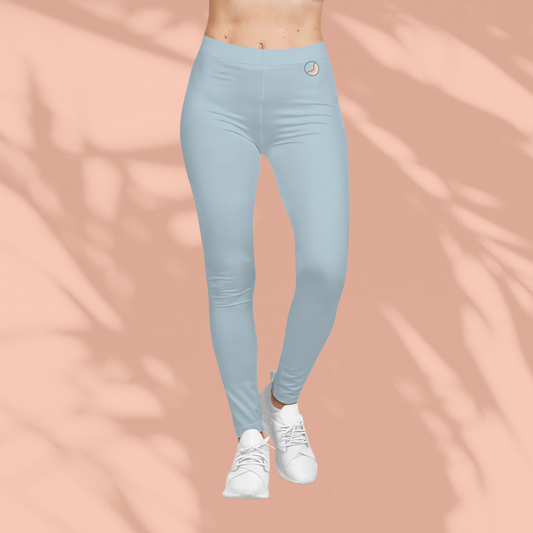 Front view of woman wearing light blue Soft & Sleek leggings with circular emblem on waistband, white sneakers, peach background