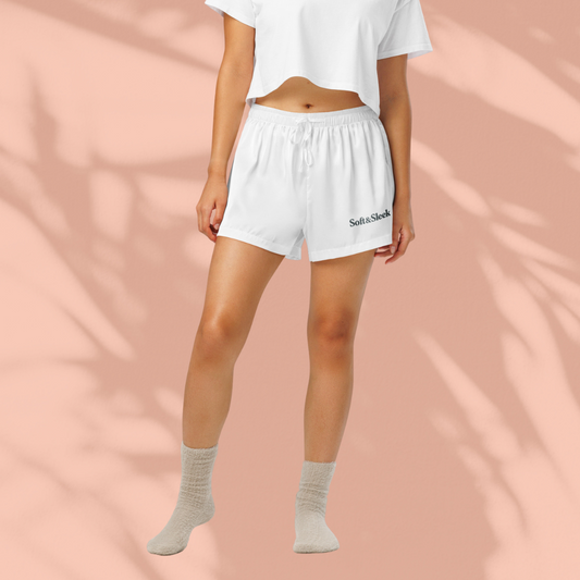 Full-body front view of a woman wearing Soft & Sleek white lounge shorts and cropped top, on a peach background with shadow overlay.
