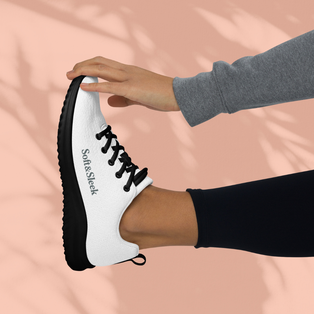 Side view of a person holding their foot up, wearing a white Soft & Sleek sneaker with black sole and black laces.