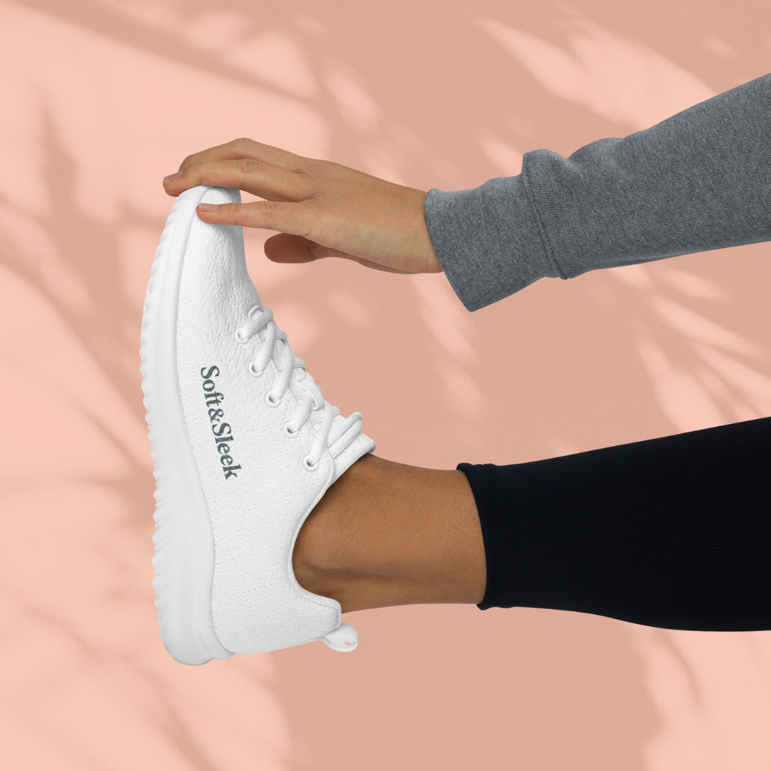 Side view of a person holding their foot up, wearing a white Soft & Sleek sneaker with the logo on the side.