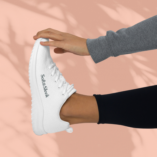 Side view of a person holding their foot up, wearing a white Soft & Sleek sneaker with the logo on the side.