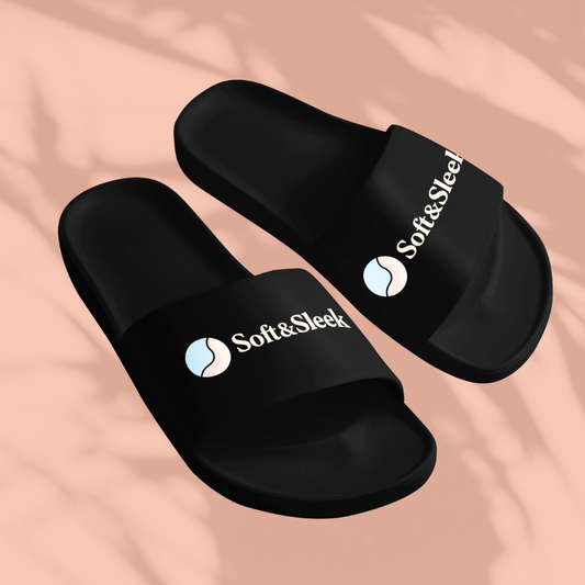 Angled top view of black Soft & Sleek slides with white logo and circular emblem, placed on peach background with soft shadows