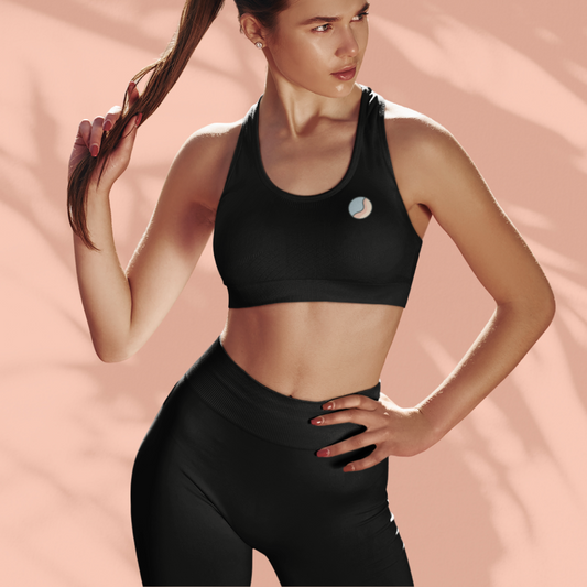 Woman wearing a black Soft & Sleek sports bra and high-waisted leggings, holding her ponytail and standing against a peach background.