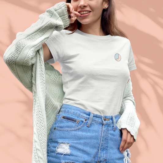 Woman wearing a white Soft & Sleek t-shirt with circular emblem on the chest, styled with a cardigan and denim shorts, peach background