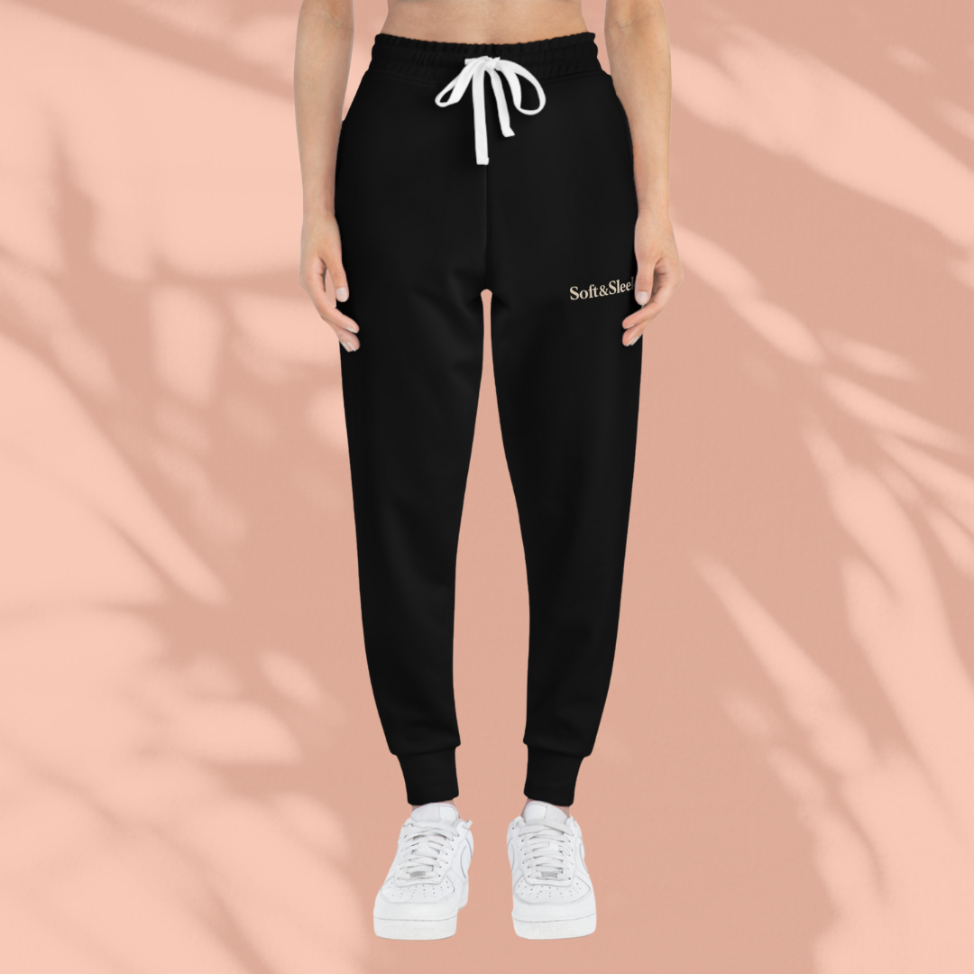 Front view of a person wearing black Soft & Sleek joggers with white drawstring and logo on left thigh, standing against a peach background.