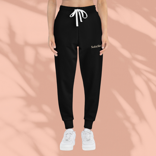 Front view of a person wearing black Soft & Sleek joggers with white drawstring and logo on left thigh, standing against a peach background.