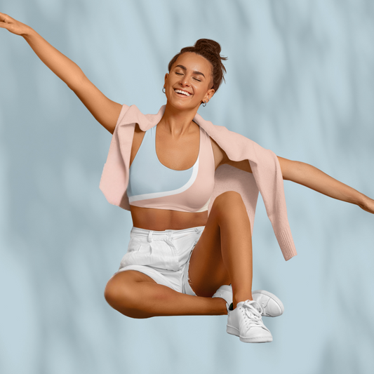 Woman wearing a multicolour Soft & Sleek sports bra with white, pastel blue, peach, and nude colour-block design, styled with white shorts and open pink jacket, pale blue background